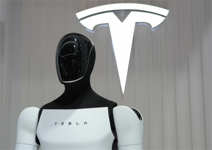 Head-and-torso shot of Tesla Optimus prototype displayed in front of illuminated Tesla brand logo at exhibition venue